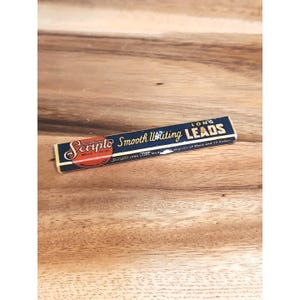 Może przedstawiać: Zabytkowe pudełko Scripto Long Leads. Pudełko jest prostokątne z granatowym tłem i złotą obwódką. Na pudełku wydrukowano napis "Scripto Smooth Writing LEADS". Pudełko ma również czerwone i złote okrągłe logo.