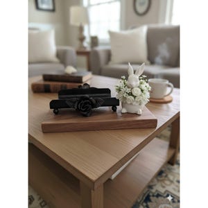 May include: A wooden tray with a black metal rose-shaped napkin holder and a white ceramic bunny vase filled with white flowers. The items are on a light brown wooden coffee table.