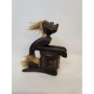 May include: A dark brown wooden sculpture of a person playing a drum. The figure has a stylized face and is adorned with natural fiber accents. The drum is cylindrical and the figure is seated.