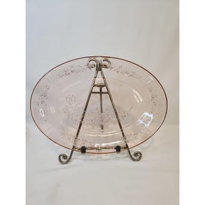 May include: A pink oval serving platter with a floral design, displayed on a decorative metal stand. The platter has a raised rim and etched floral patterns. The stand is made of dark metal with decorative scrollwork.