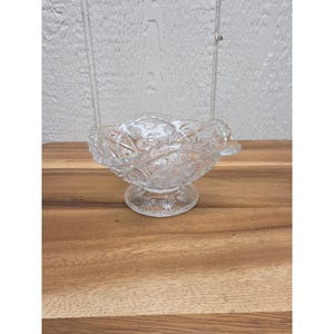 May include: A clear, vintage glass bowl with a pedestal base. The bowl features a detailed, embossed pattern of stars and floral designs. The bowl is likely used for serving or display purposes. The bowl is sitting on a wooden surface.