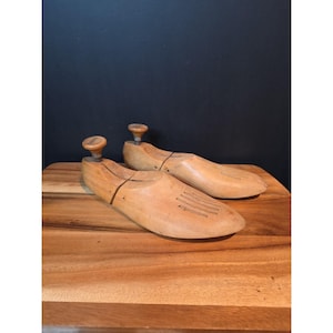 May include: Two wooden shoe stretchers with adjustable knobs. The shoe forms are light brown with a natural wood grain. The stretchers are on a wooden surface with a dark background.