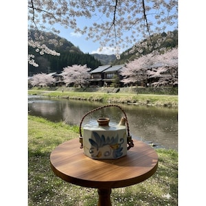 Puede incluir: Una tetera de cerámica con un asa trenzada marrón y diseños florales azules y amarillos descansa sobre una pequeña mesa de madera. El fondo presenta un río, edificios tradicionales y cerezos en flor.