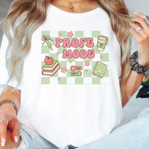 May include: A white t-shirt featuring a light green and white checkered graphic with the text PROFE MODE in pink. The design includes illustrations of a pencil, a coffee cup, a red apple on books, a toggle switch, and a notebook with small flowers.