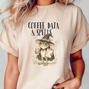 Könnte beinhalten: Ein beiges T-Shirt mit einer Grafik eines grünen Frosches, der einen dunklen spitzen Hut und einen Umhang trägt. Der Frosch hält einen Kaffeebecher und ein Klemmbrett. Über dem Frosch steht der Text COFFEE, DATA & SPELLS in schwarzer Schrift.