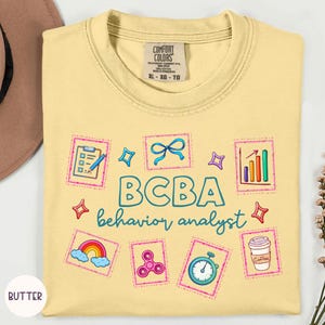 May include: A pale yellow t-shirt featuring the text BCBA behavior analyst in blue. The design includes colorful illustrations of a clipboard, a blue bow, a bar graph, a rainbow, a pink fidget spinner, a stopwatch, and a coffee cup.