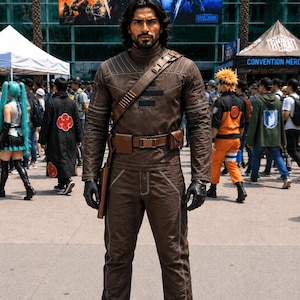May include: A person in a brown costume with a high collar, shoulder strap, and belt. The costume includes a jacket, pants, and boots. The person is wearing black gloves. The background shows a crowd of people and convention banners.