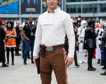 May include: A long-sleeved white shirt with a high collar and a diagonal front flap, paired with brown trousers featuring a vertical gold stripe along the side, a dark brown leather belt with a metallic buckle, and a brown leather holster attached.