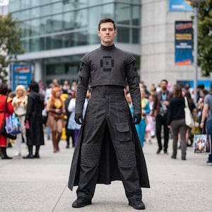 K&ouml;nnte beinhalten: Eine Person tr&auml;gt ein dunkelgraues Kost&uuml;m mit hohem Kragen, kurzer Jacke und langer Hose mit Knieschonern. Das Kost&uuml;m hat ein geometrisches Design auf der Brust und wird mit schwarzen Handschuhen und Schuhen kombiniert. Die Person steht in einer Menschenmenge.