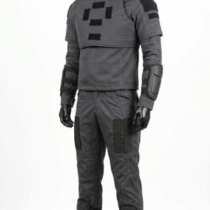 Pu&ograve; includere: Un set di uniforme tattica grigio scuro su un manichino, composto da una maglia a maniche lunghe con collo alto e pettorina, pantaloni abbinati con ginocchiere rinforzate, protezioni per gli avambracci e stivali neri.