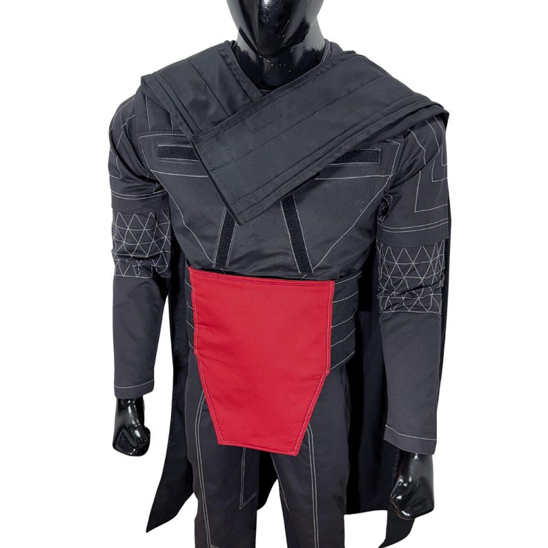 Puede incluir: Traje negro con un panel rojo en la cintura. El disfraz tiene costuras blancas y patrones geom&eacute;tricos en las mangas. El maniqu&iacute; lleva una capa y guantes negros.