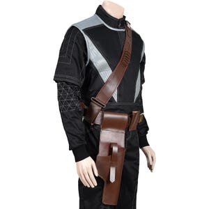 May include: A brown leather holster and belt set with a shoulder strap. The holster is designed to hold a weapon. The belt has pouches and a buckle. The set is worn over a black jumpsuit with gray accents.