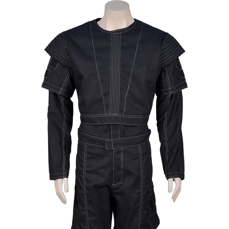 May include: Black jumpsuit with white stitching details. The outfit features long sleeves, a round neckline, and a belt at the waist. The shoulders have layered accents, and the shorts have cargo pockets.