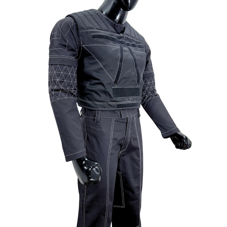 May include: Black tactical jacket and pants set. The jacket features a quilted design on the shoulders and chest, with white stitching outlining the panels and geometric patterns on the sleeves. The pants have white stitching along the seams.