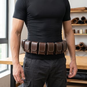 May include: A brown leather utility belt featuring multiple rectangular pouches with flap closures and visible white stitching. The belt is worn around the waist, consisting of several individual compartments arranged side by side in a continuous row.