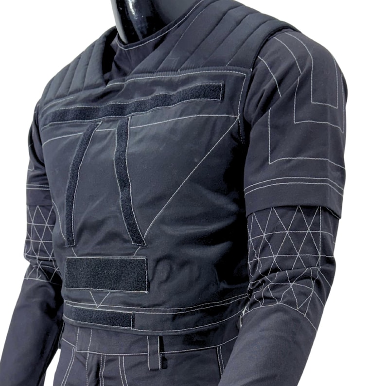 May include: Black quilted jacket with white stitching and velcro closures. The jacket features a geometric design on the sleeves and chest. The garment is displayed on a mannequin.