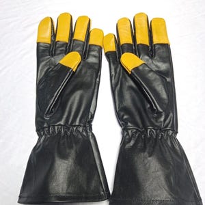 May include: A pair of black leather gloves featuring long cuffs and contrasting yellow leather fingertips. The gloves have a smooth finish, stitched seams, and a gathered elastic wrist section to ensure a secure fit during use.