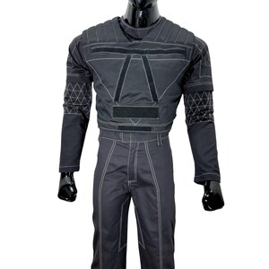 May include: Black jumpsuit with white stitching and geometric patterns on the sleeves. The top features a padded design with velcro closures and a crew neck. The pants have a straight leg cut. The outfit is displayed on a black mannequin.
