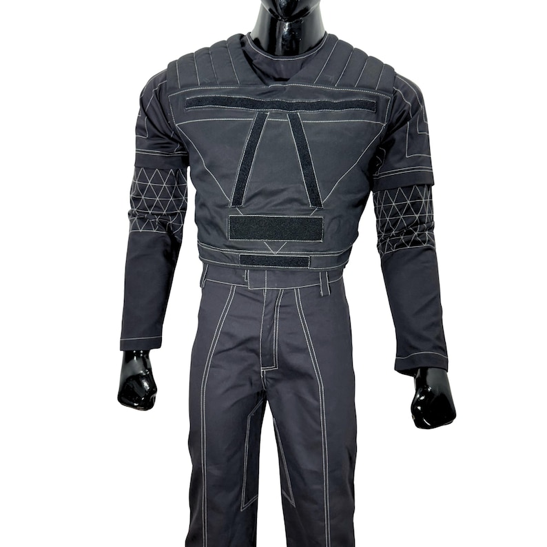 May include: Black jumpsuit with white stitching and geometric patterns on the sleeves. The top features a padded design with velcro closures and a crew neck. The pants have a straight leg cut. The outfit is displayed on a black mannequin.