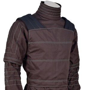 Puede incluir: Chaqueta protectora marr&oacute;n con cuello alto y hombreras. La chaqueta presenta detalles de costuras blancas y un dise&ntilde;o de manga en capas. El dise&ntilde;o general sugiere un enfoque en la durabilidad y la protecci&oacute;n.