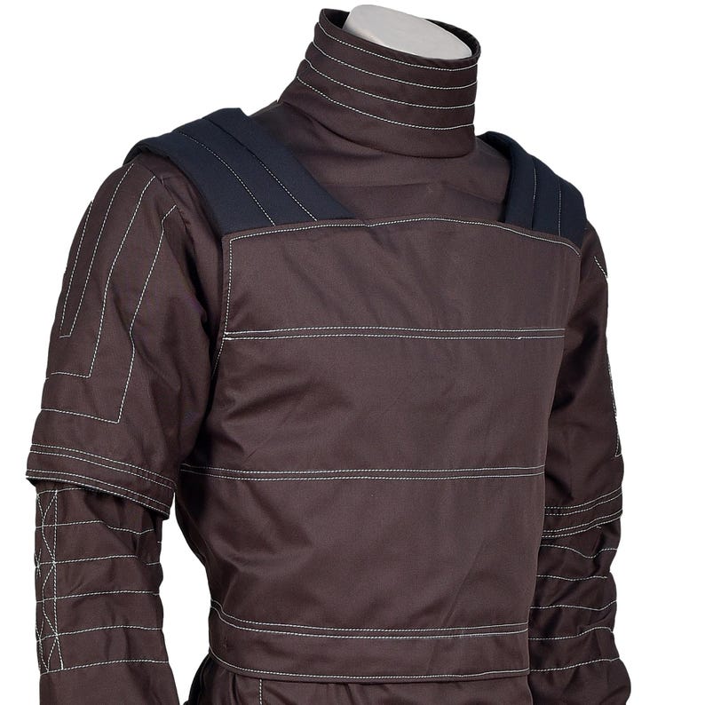 Puede incluir: Chaqueta protectora marr&oacute;n con cuello alto y hombreras. La chaqueta presenta detalles de costuras blancas y un dise&ntilde;o de manga en capas. El dise&ntilde;o general sugiere un enfoque en la durabilidad y la protecci&oacute;n.