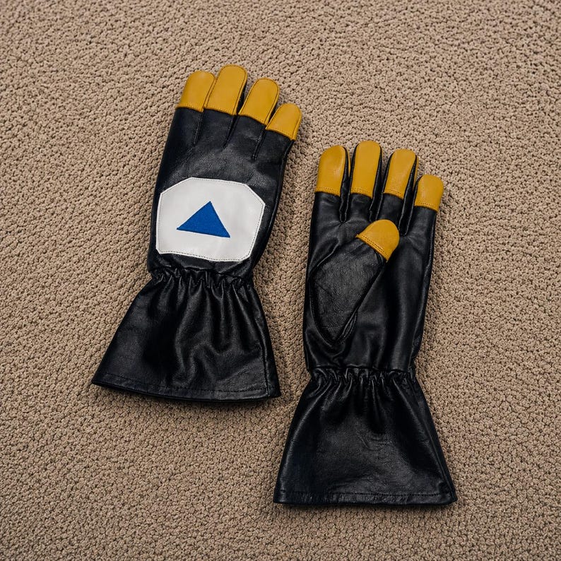May include: A pair of black gloves featuring yellow fingertips, a flared wrist cuff, and a white patch on the back of one glove displaying a blue triangle. The gloves are constructed with a smooth finish and include an elasticized wrist section.