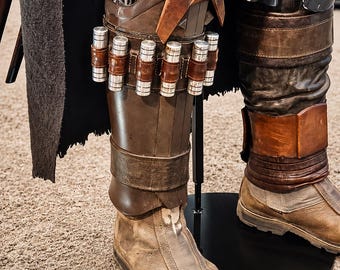 May include: A costume leg armor set featuring dark brown molded shin guards with a leather strap holding six silver cylindrical canisters. The armor includes weathered tan boots, a dark grey textured cape, and metallic knee plates with scuff marks.