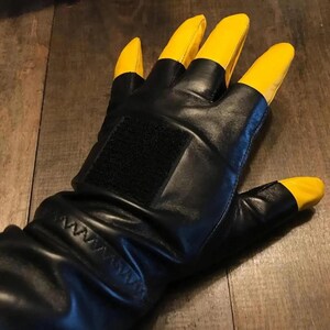 May include: A black leather glove featuring bright yellow fingertips and a thumb tip. The glove includes a rectangular black hook-and-loop fastener patch on the back of the hand and a stitched cuff detail near the wrist area for a secure fit.