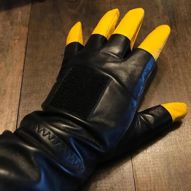 May include: A black leather glove featuring bright yellow fingertips and a thumb tip. The glove includes a rectangular black hook-and-loop fastener patch on the back of the hand and a stitched cuff detail near the wrist area for a secure fit.