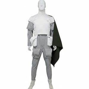 May include: A futuristic costume displayed on a mannequin. The outfit includes a white top, gray pants with pockets, and a dark green cape. The mannequin wears white boots. The costume is designed with a high collar.