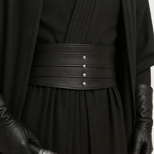 May include: A black leather belt with four horizontal bands and silver-colored rivets. The belt is worn over a black outfit, with black gloves visible on the sides. The belt is a fashion accessory.