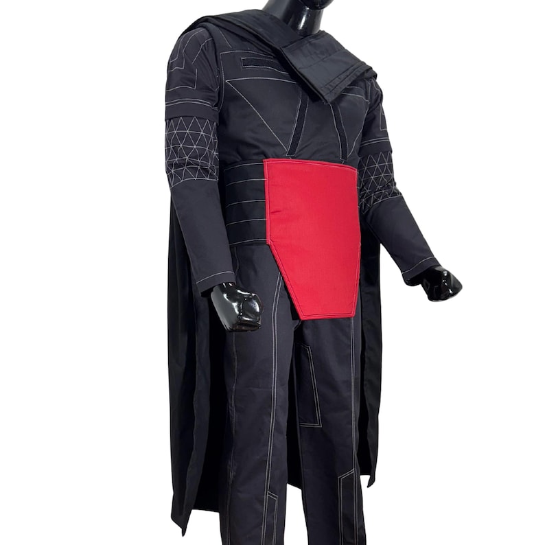 Puede incluir: Traje negro con secci&oacute;n media roja y capa. El disfraz presenta detalles de costura blancos y patrones geom&eacute;tricos en las mangas. El atuendo se muestra en un maniqu&iacute;.