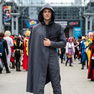 Könnte beinhalten: Eine Person trägt einen dunkelgrauen Kapuzenmantel und eine passende Hose. Der Umhang hat ein kapuzenartiges Design und ein geometrisches Muster auf der Schulter. Die Person steht in einer Menschenmenge, wahrscheinlich auf einer Convention oder Veranstaltung.