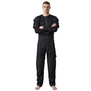 May include: Black jumpsuit with white stitching details. The one-piece garment features a round neck, long sleeves, and multiple pockets. The pants have reinforced knee panels. The model is standing with arms crossed.