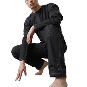 May include: Black jumpsuit with white stitching details. The long-sleeved top and pants feature visible seams and reinforced patches. The model is squatting, showcasing the garment's design and fit. The jumpsuit is a modern take on workwear.