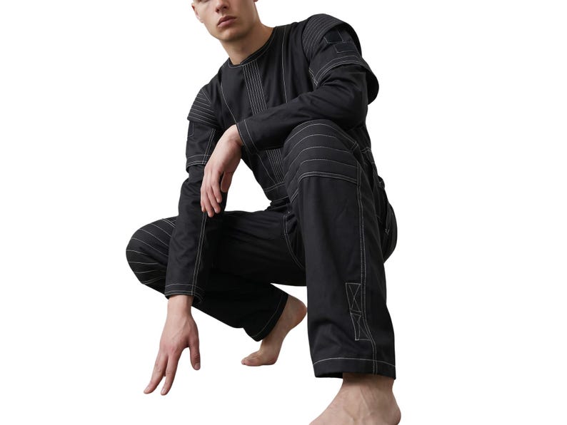 May include: Black jumpsuit with white stitching details. The long-sleeved top and pants feature visible seams and reinforced patches. The model is squatting, showcasing the garment's design and fit. The jumpsuit is a modern take on workwear.