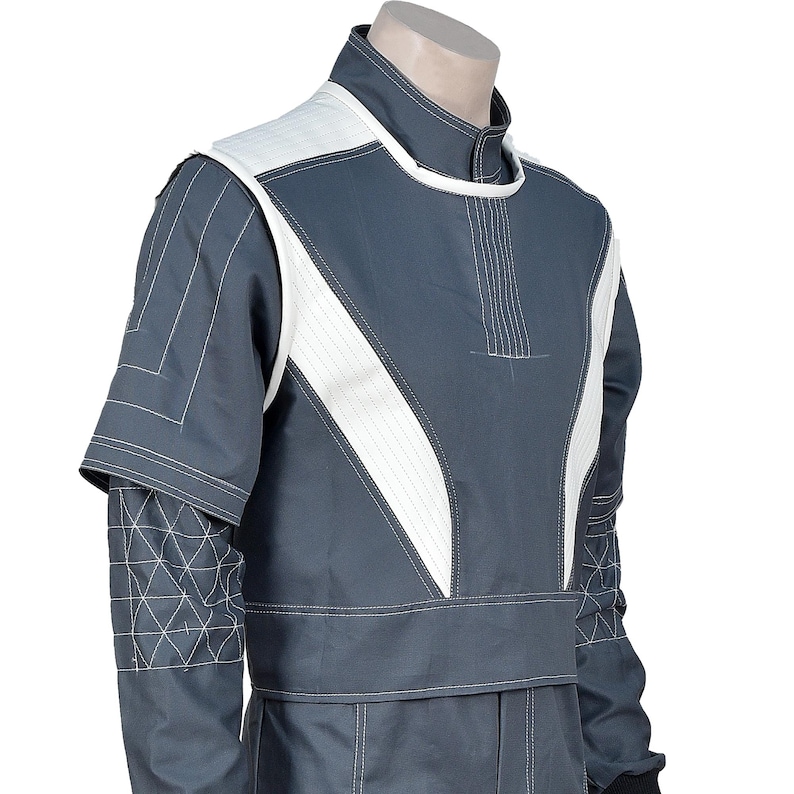 May include: A gray jumpsuit with white accents and stitching. The upper portion features a white panel across the chest and shoulders, with a high collar. The sleeves have a geometric pattern on the lower arm. The jumpsuit appears to be made of a durable material.