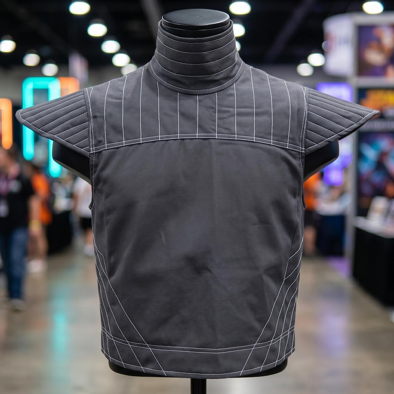 Op de afbeelding: Een grijze vest met witte stiksels en een hoge kraag, tentoongesteld op een mannequin. De vest heeft een gewatteerd ontwerp op de schouders en verticale lijnen op de borst. De algehele stijl suggereert een futuristisch of cosplay thema.