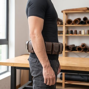May include: A brown leather utility belt featuring multiple rectangular pouches with flap closures and visible white stitching. The belt is worn around the waist, showcasing a structured design with individual compartments for storage and organisation.