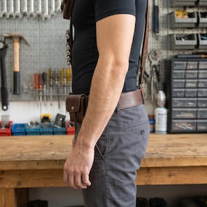 May include: A brown leather shoulder harness and belt set featuring a small rectangular pouch on the chest, a vertical row of five cylindrical loops for holding items, and a matching pouch attached to the waist belt, worn over a black t-shirt.