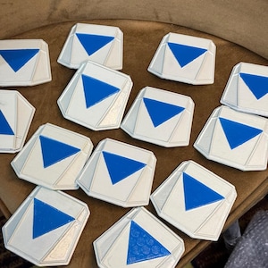 May include: A collection of twelve white, multi-faceted geometric tokens, each featuring a centered, downward-pointing blue triangle. The tokens have a raised, angular border design and a smooth, matte finish, arranged in a scattered cluster.