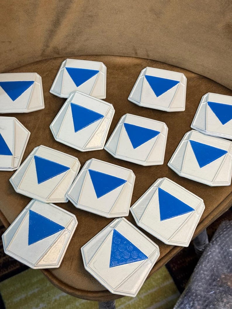 May include: A collection of twelve white, multi-faceted geometric tokens, each featuring a centered, downward-pointing blue triangle. The tokens have a raised, angular border design and a smooth, matte finish, arranged in a scattered cluster.
