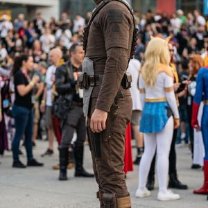 May include: A person in a brown costume with a high collar, shoulder armor, and a utility belt. The outfit includes matching pants and boots. The person is standing in a crowd of people, likely at a convention.