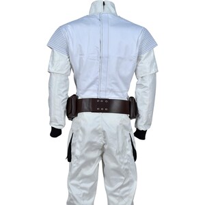 May include: A white jumpsuit with a high collar and a brown belt with two pouches. The back of the jacket has a structured design with horizontal lines on the shoulders. Black cuffs are visible at the wrists.