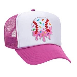 May include: A baseball cap with a white front panel and a pink brim and mesh back. The cap features an embroidered design of a baseball with sprinkles and dripping pink icing. The cap is a trucker style.