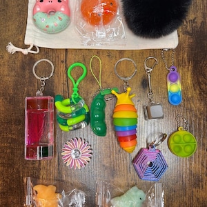 May include: A selection of colourful keychains and fidget toys, including a pink donut, orange ball, black pom-pom, and various keychains with charms like a rainbow snail, pea pod, and a hexagon. The items are arranged on a wooden surface.