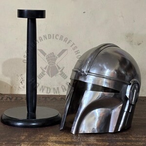 May include: A metallic silver Mandalorian helmet with a visor, resting on a wooden surface. A black helmet stand is to the left. The helmet has a polished finish and is a replica of the iconic character's headgear.