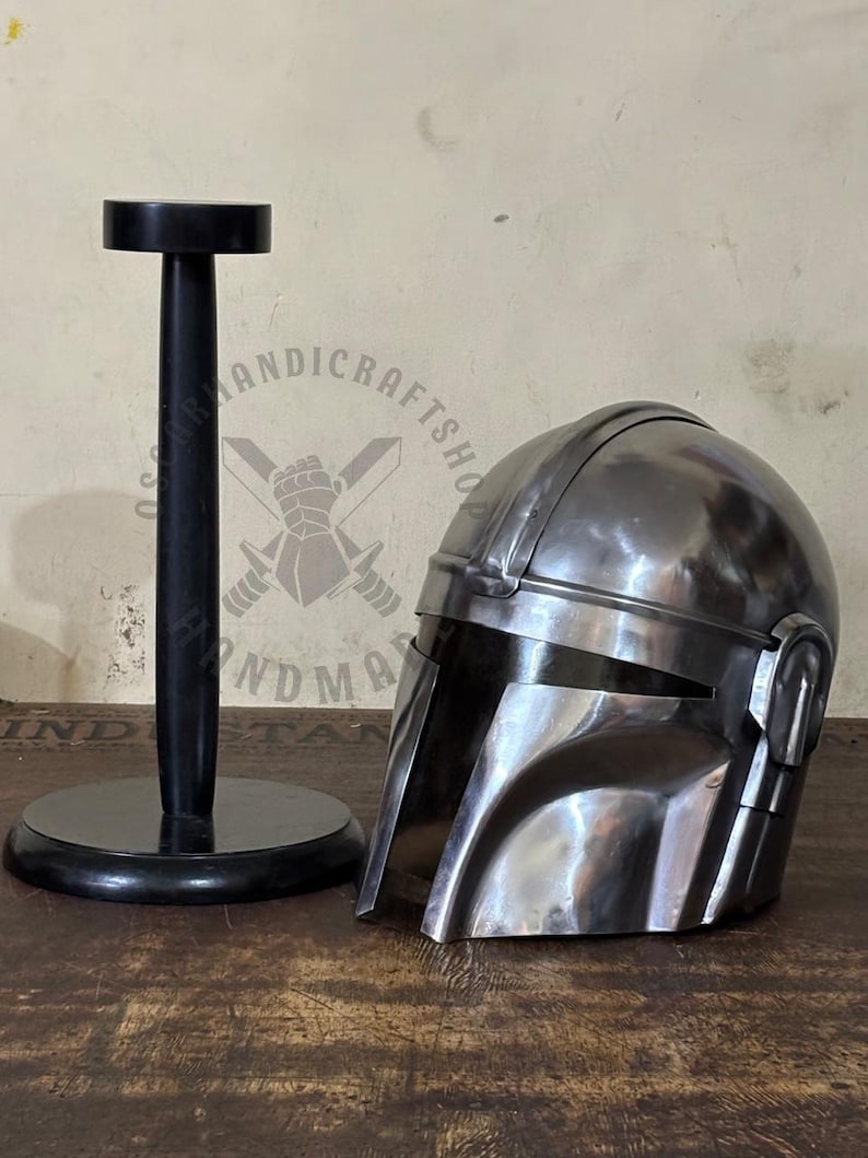 May include: A metallic silver Mandalorian helmet with a visor, resting on a wooden surface. A black helmet stand is to the left. The helmet has a polished finish and is a replica of the iconic character's headgear.