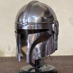 May include: A metallic silver helmet with a visor and a rounded top, displayed on a black stand. The helmet has a detailed design with visible seams and a polished finish. The base has the text "SCARHANDIC SHOP HAND MADE".