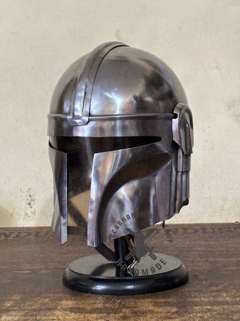 May include: A metallic silver helmet with a visor and a rounded top, displayed on a black stand. The helmet has a detailed design with visible seams and a polished finish. The base has the text "SCARHANDIC SHOP HAND MADE".